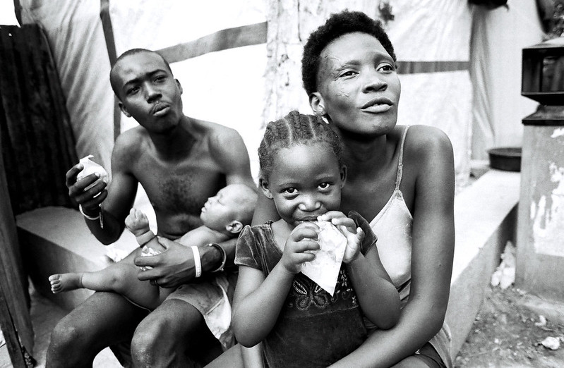 Black-and-white photograph of a family on a Port au Prince street: a woman holding a young girl, with a man and an infant beside them.
