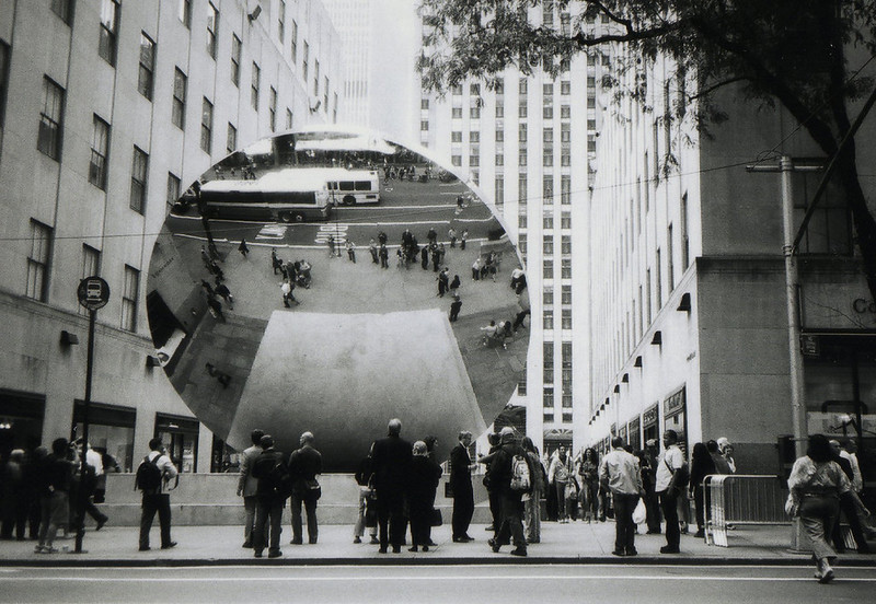 A black-and-white photo of a massive polished steel orb sculpture on a Midtown Manhattan street, reflecting an upside-down view of the surrounding buildings and pedestrians. A crowd of onlookers lines the sidewalk below it.