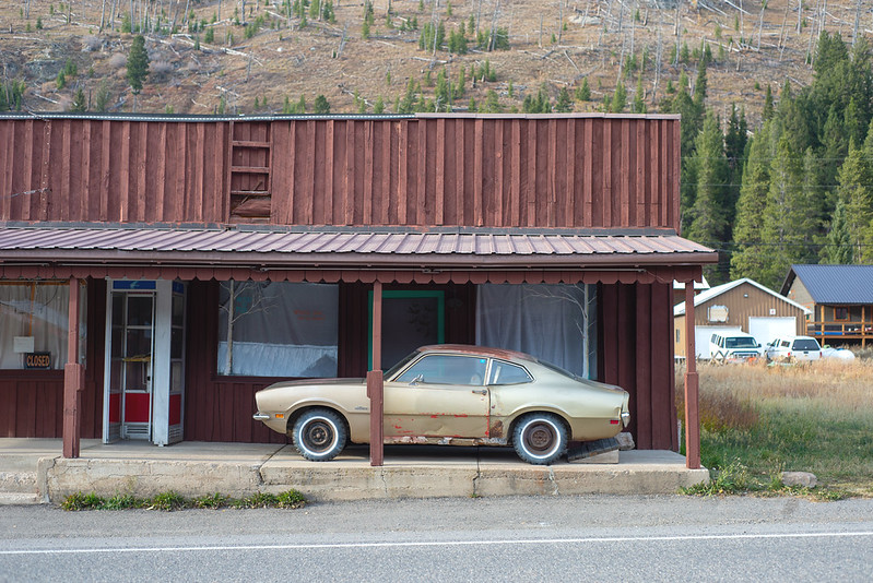 A closed storefront in a small mountain town, with an old gold Ford Maverick parked under the porch and pine-covered slopes rising behind it.
