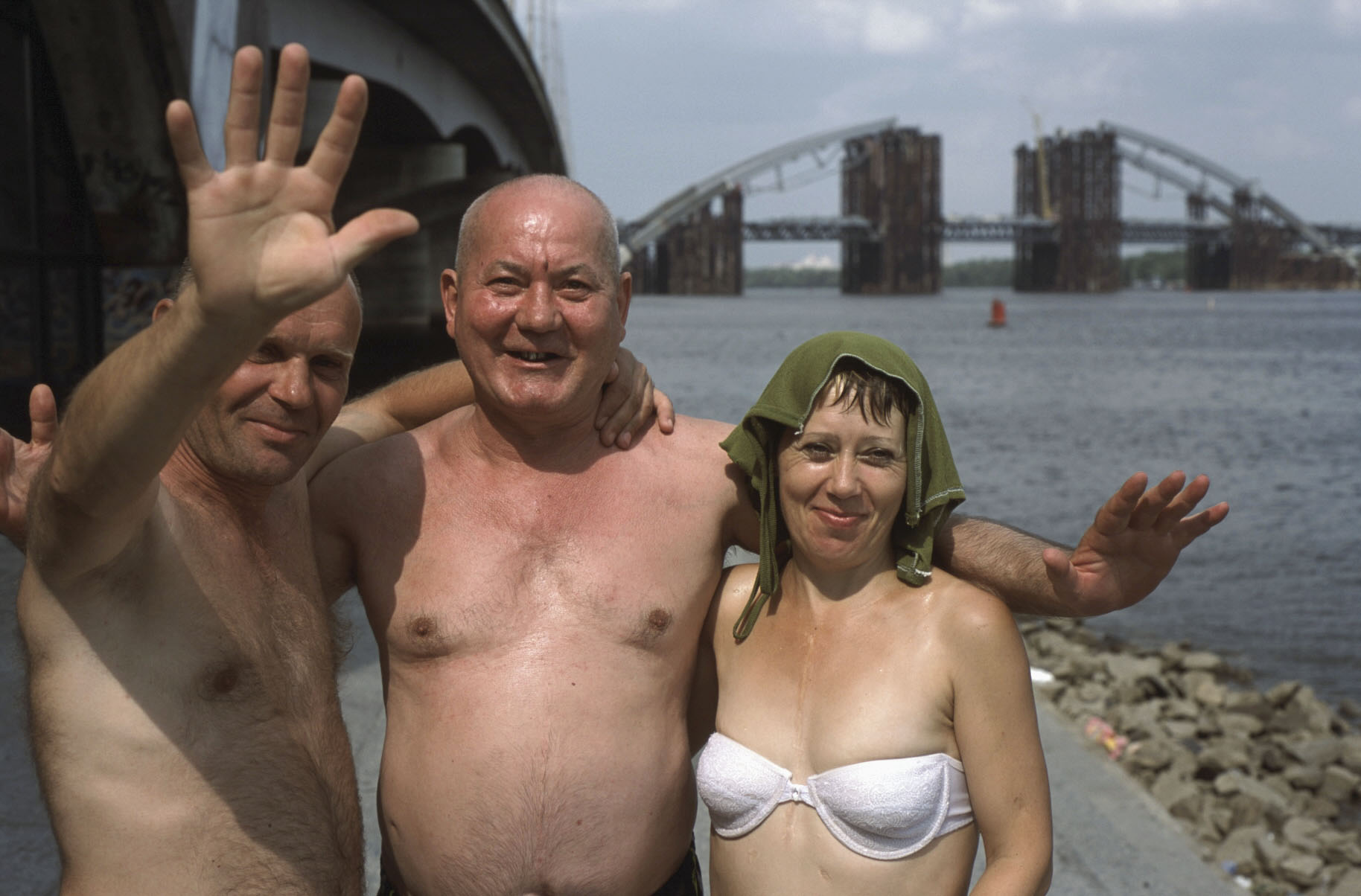 Three smiling sunbathers, two shirtless men and a woman in a white bikini top with a green cloth draped over her head, pose by a river. Two of them wave, with a half-built steel arch bridge across the water behind them.