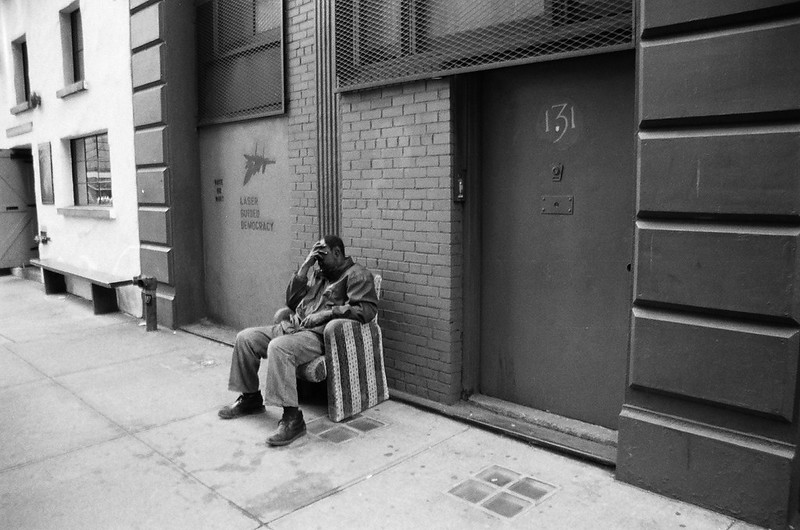Black-and-white street photograph of a man seated against a brick building, one hand to his face, beside stenciled graffiti reading 'LASER GUIDED DEMOCRACY' with a fighter-jet silhouette.