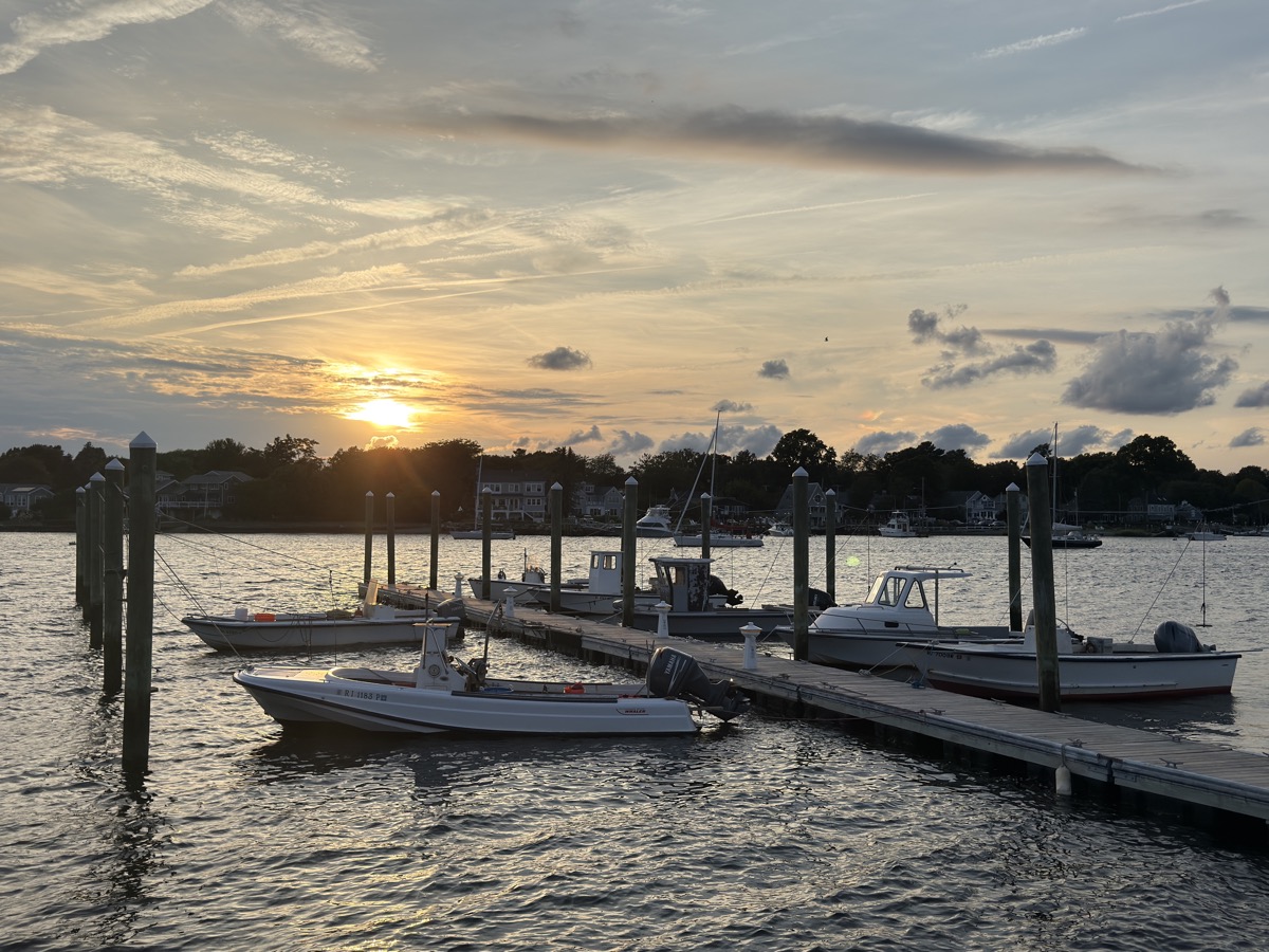 Small fishing and motor boats tied along a wooden dock in a calm harbor at sunset, with houses and trees on the far shore.