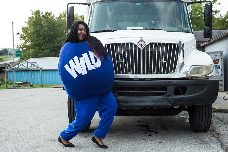A person in a large round blue 'WILD' mascot costume strikes a pose in front of a white box truck in a gravel lot.