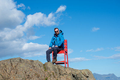 Steve sitting on a red chair on a rocky summit under a big blue sky.